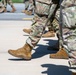 The image shows a close-up view of military personnel marching in formation on a paved surface. Multiple soldiers are visible from approximately the waist down, wearing matching camouflage uniforms and tan combat boots. The tight framing emphasizes the uniformity and coordinated movement of the group.