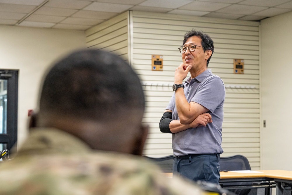 A civilian man wearing glasses and a gray polo shirt stands at the front of what appears to be a military classroom or briefing room, engaged in a thoughtful pose with arms crossed and one hand raised to his chin. In the foreground, the back of a person in military camouflage uniform is visible, suggesting an audience of military personnel. The setting appears to be an indoor institutional facility with drop ceilings and wall-mounted storage units.