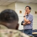 A civilian man wearing glasses and a gray polo shirt stands at the front of what appears to be a military classroom or briefing room, engaged in a thoughtful pose with arms crossed and one hand raised to his chin. In the foreground, the back of a person in military camouflage uniform is visible, suggesting an audience of military personnel. The setting appears to be an indoor institutional facility with drop ceilings and wall-mounted storage units.
