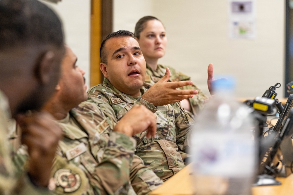 Military personnel in OCP (Operational Camouflage Pattern) uniforms are seated at a table during what appears to be a briefing, meeting, or training session. A male service member in the center is actively speaking and gesturing with his hand, engaging with others at the table. At least three uniformed individuals are visible, with equipment and a water bottle on the table.