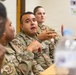 Military personnel in OCP (Operational Camouflage Pattern) uniforms are seated at a table during what appears to be a briefing, meeting, or training session. A male service member in the center is actively speaking and gesturing with his hand, engaging with others at the table. At least three uniformed individuals are visible, with equipment and a water bottle on the table.