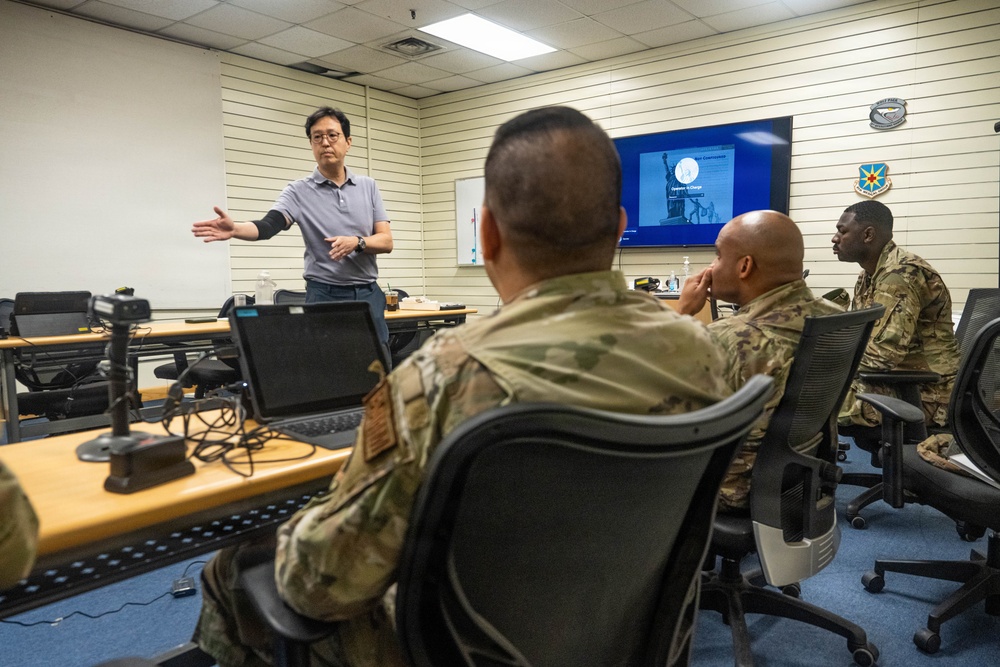 A civilian instructor is delivering a briefing or training session to a group of military personnel in camouflage uniforms inside a conference or classroom setting. A presentation slide is visible on a large monitor in the background. The setting appears to be a military installation based on the unit insignia patches visible on the wall.