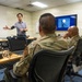 A civilian instructor is delivering a briefing or training session to a group of military personnel in camouflage uniforms inside a conference or classroom setting. A presentation slide is visible on a large monitor in the background. The setting appears to be a military installation based on the unit insignia patches visible on the wall.