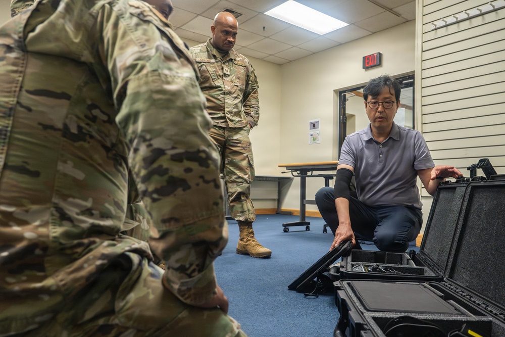 A civilian in a gray polo shirt is demonstrating or presenting equipment from a hard-shell tactical case to two U.S. Army soldiers in OCP (Operational Camouflage Pattern) uniforms. The setting appears to be an indoor briefing or training room on a military installation. The interaction suggests a product demonstration, equipment familiarization, or technical briefing.