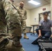 A civilian in a gray polo shirt is demonstrating or presenting equipment from a hard-shell tactical case to two U.S. Army soldiers in OCP (Operational Camouflage Pattern) uniforms. The setting appears to be an indoor briefing or training room on a military installation. The interaction suggests a product demonstration, equipment familiarization, or technical briefing.