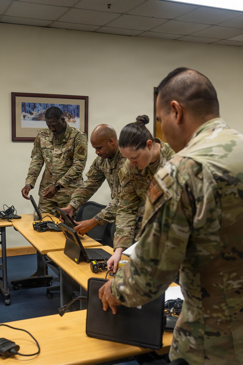 Four military personnel in OCP (Operational Camouflage Pattern) uniforms are gathered around a wooden table in an office or classroom setting, working with laptops and tablet devices. They appear to be setting up or configuring electronic equipment, possibly during a training or operational briefing session. A framed photograph of a wolf in a winter scene is visible on the wall behind them.