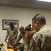 Four military personnel in OCP (Operational Camouflage Pattern) uniforms are gathered around a wooden table in an office or classroom setting, working with laptops and tablet devices. They appear to be setting up or configuring electronic equipment, possibly during a training or operational briefing session. A framed photograph of a wolf in a winter scene is visible on the wall behind them.