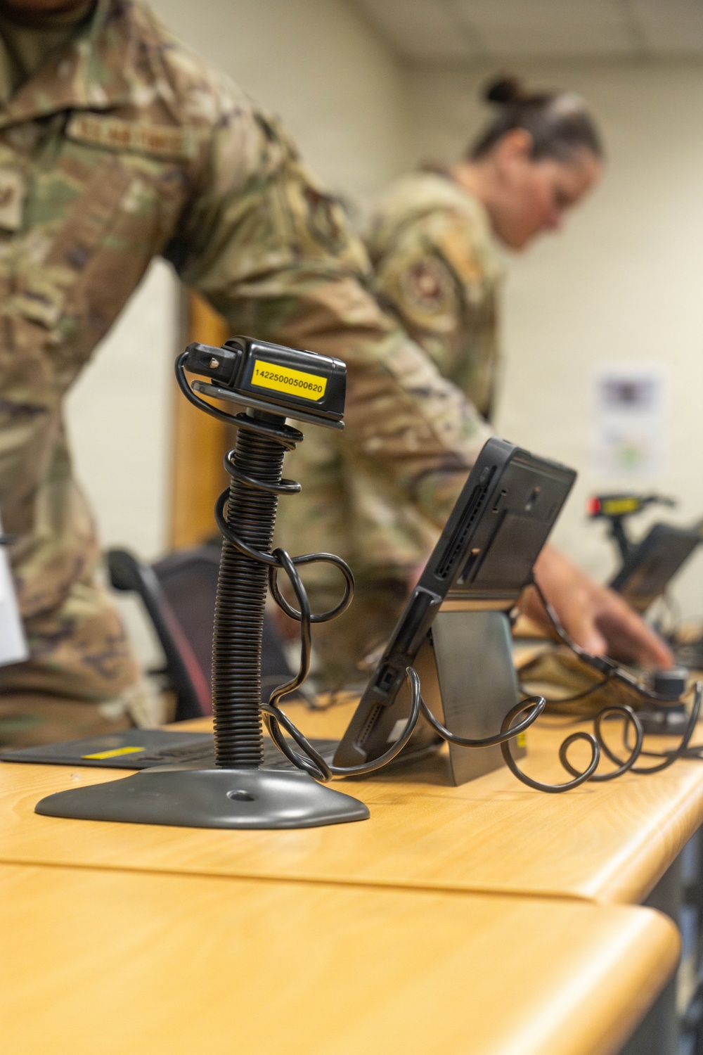 Two military personnel in OCP (Operational Camouflage Pattern) uniforms are working at a desk equipped with scanning and processing equipment. The foreground features a barcode scanner on a coiled-cord pedestal stand with a visible asset tag numbered '14225000500620'. The setting appears to be a military administrative or logistics processing station.