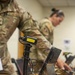 Two military personnel in OCP (Operational Camouflage Pattern) uniforms are working at a desk equipped with scanning and processing equipment. The foreground features a barcode scanner on a coiled-cord pedestal stand with a visible asset tag numbered '14225000500620'. The setting appears to be a military administrative or logistics processing station.