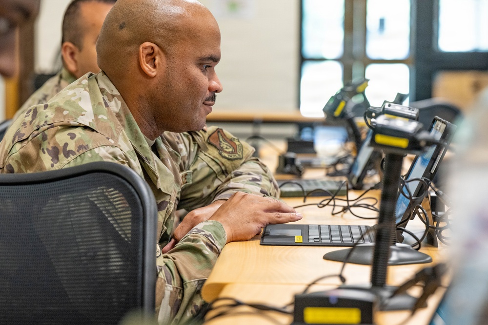 A military service member in OCP (Operational Camouflage Pattern) uniform is seated at a workstation, focused on a computer screen. The setting appears to be a military operations center or training facility with multiple workstations visible. A second uniformed individual is partially visible in the background.