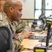 A military service member in OCP (Operational Camouflage Pattern) uniform is seated at a workstation, focused on a computer screen. The setting appears to be a military operations center or training facility with multiple workstations visible. A second uniformed individual is partially visible in the background.