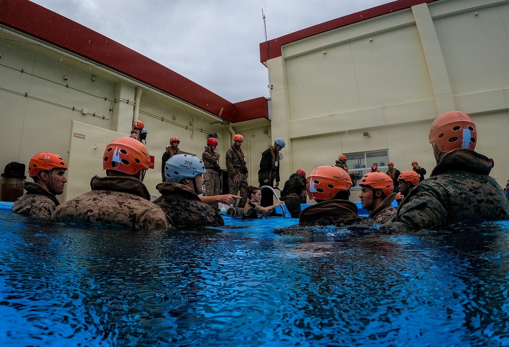 A group of military personnel, appearing to be U.S. Marines based on their camouflage uniforms, are participating in a water survival or aquatic training exercise in an outdoor pool. Most participants are submerged to chest level and wearing orange safety helmets, while an instructor in a white helmet appears to be giving directions. The training takes place adjacent to a military-style building under overcast, rainy conditions.