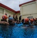 A group of military personnel, appearing to be U.S. Marines based on their camouflage uniforms, are participating in a water survival or aquatic training exercise in an outdoor pool. Most participants are submerged to chest level and wearing orange safety helmets, while an instructor in a white helmet appears to be giving directions. The training takes place adjacent to a military-style building under overcast, rainy conditions.