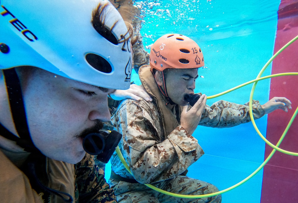 Two individuals in military camouflage uniforms are submerged underwater during what appears to be a water survival or combat swimmer training exercise. Both wear Pro-Tec helmets — one white and one orange numbered '15' — and are breathing through air hoses or mouthpieces. The underwater setting, safety equipment, and structured positioning suggest a controlled military training environment.