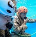 Two individuals in military camouflage uniforms are submerged underwater during what appears to be a water survival or combat swimmer training exercise. Both wear Pro-Tec helmets — one white and one orange numbered '15' — and are breathing through air hoses or mouthpieces. The underwater setting, safety equipment, and structured positioning suggest a controlled military training environment.