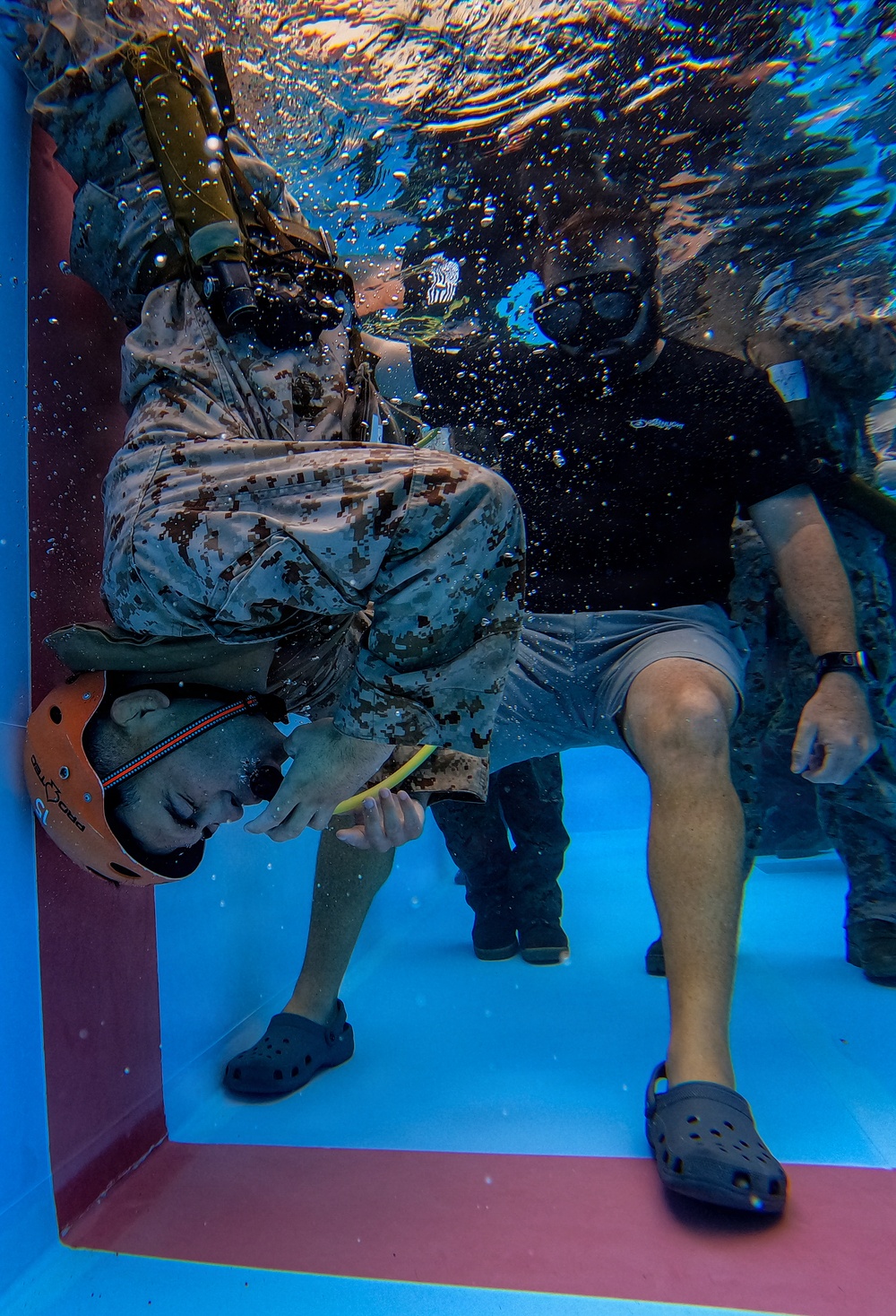 An underwater photograph showing what appears to be a military water survival or egress training exercise. A person wearing MARPAT (Marine digital camouflage) uniform and tactical gear is submerged and bent forward in a doorframe-like structure, while an instructor wearing a black shirt, dive mask, and Crocs sandals observes nearby. The image captures air bubbles rising to the surface, indicating active movement and exhalation underwater.
