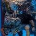 An underwater photograph showing what appears to be a military water survival or egress training exercise. A person wearing MARPAT (Marine digital camouflage) uniform and tactical gear is submerged and bent forward in a doorframe-like structure, while an instructor wearing a black shirt, dive mask, and Crocs sandals observes nearby. The image captures air bubbles rising to the surface, indicating active movement and exhalation underwater.