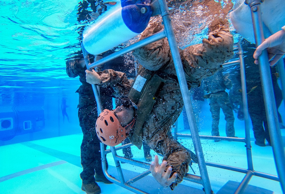 An underwater photograph captures military personnel conducting what appears to be a helicopter underwater egress training (HUET) exercise in a pool facility. A trainee wearing a pink/orange safety helmet and military camouflage uniform is navigating through a submerged metal cage structure, simulating escape from a downed aircraft. Multiple other uniformed personnel are visible assisting or observing the exercise underwater.