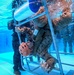 An underwater photograph captures military personnel conducting what appears to be a helicopter underwater egress training (HUET) exercise in a pool facility. A trainee wearing a pink/orange safety helmet and military camouflage uniform is navigating through a submerged metal cage structure, simulating escape from a downed aircraft. Multiple other uniformed personnel are visible assisting or observing the exercise underwater.
