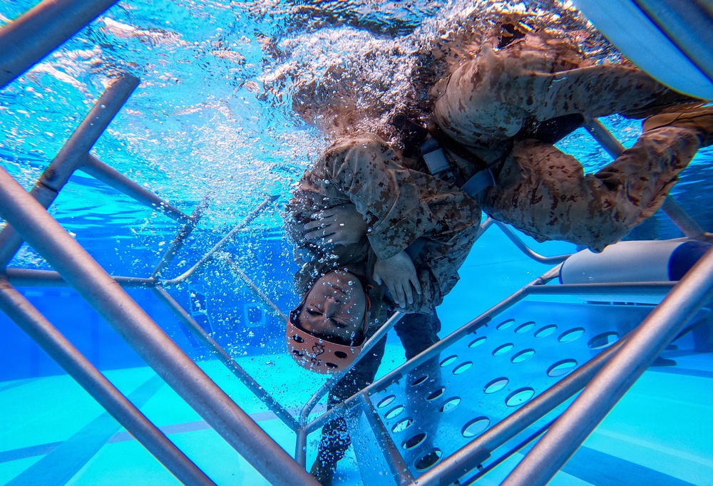 An underwater photograph captures military personnel in camouflage uniforms submerged in a pool, appearing to conduct water survival or egress training. The image is taken from below, showing individuals navigating through a metal cage or frame structure underwater. Bubbles and water turbulence indicate active movement, consistent with a controlled training exercise.