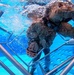 An underwater photograph captures military personnel in camouflage uniforms submerged in a pool, appearing to conduct water survival or egress training. The image is taken from below, showing individuals navigating through a metal cage or frame structure underwater. Bubbles and water turbulence indicate active movement, consistent with a controlled training exercise.
