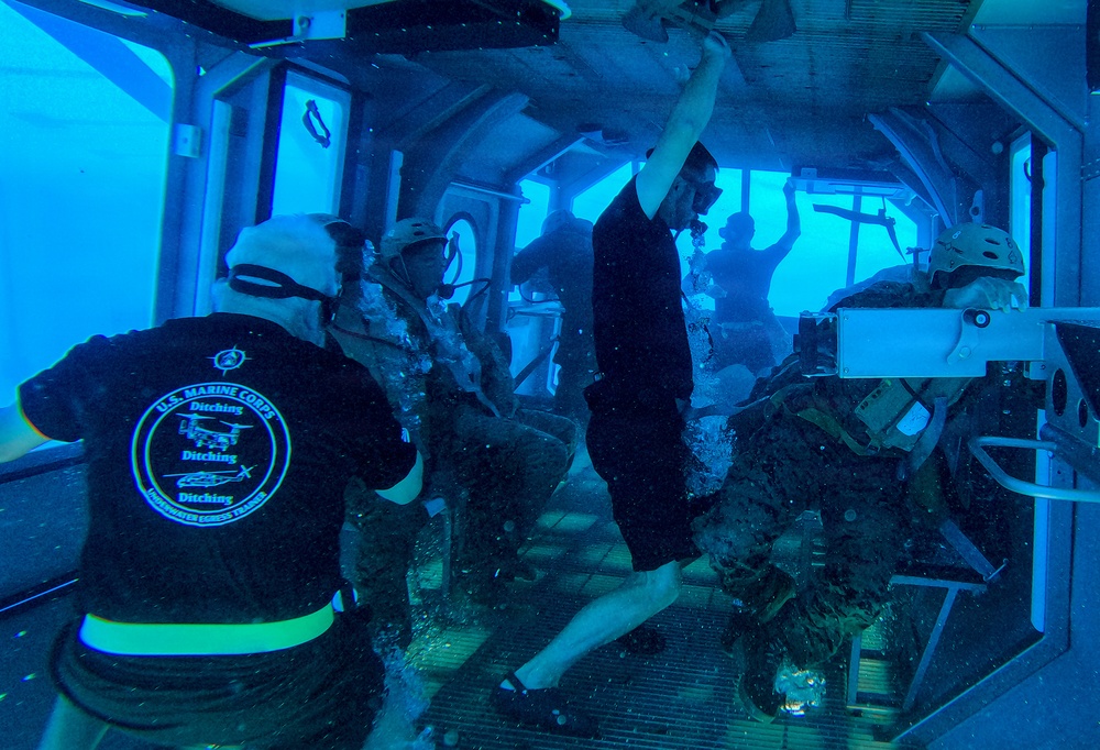 This image depicts a U.S. Marine Corps Underwater Egress Training exercise taking place inside a submerged simulator. Multiple personnel in military gear and helmets are practicing emergency escape procedures from a simulated aircraft or vehicle cabin underwater. An instructor wearing a black U.S. Marine Corps 'Ditching - Underwater Egress Trainer' shirt is visible supervising the exercise.
