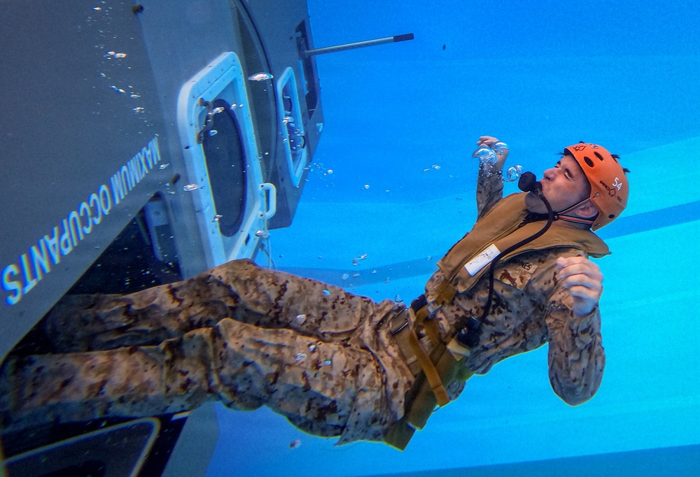 An underwater photograph shows a military personnel in desert camouflage uniform conducting a water survival or egress training exercise. The individual is submerged next to what appears to be a helicopter or aircraft fuselage simulator, wearing an orange safety helmet and an inflatable life vest. Air bubbles are visible around the person, indicating active exhalation underwater.