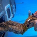 An underwater photograph shows a military personnel in desert camouflage uniform conducting a water survival or egress training exercise. The individual is submerged next to what appears to be a helicopter or aircraft fuselage simulator, wearing an orange safety helmet and an inflatable life vest. Air bubbles are visible around the person, indicating active exhalation underwater.