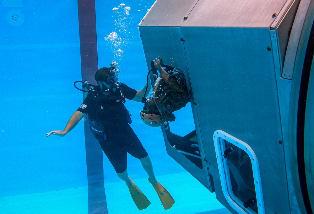 An underwater photograph taken in a training pool shows a scuba diver in a black wetsuit with yellow fins assisting a second person who is exiting or entering through a window or hatch of a submerged simulated aircraft or vehicle fuselage. The scene appears to be a military or emergency survival training exercise, likely helicopter underwater escape training (HUET). Air bubbles are visible rising from the diver's regulator toward the surface.