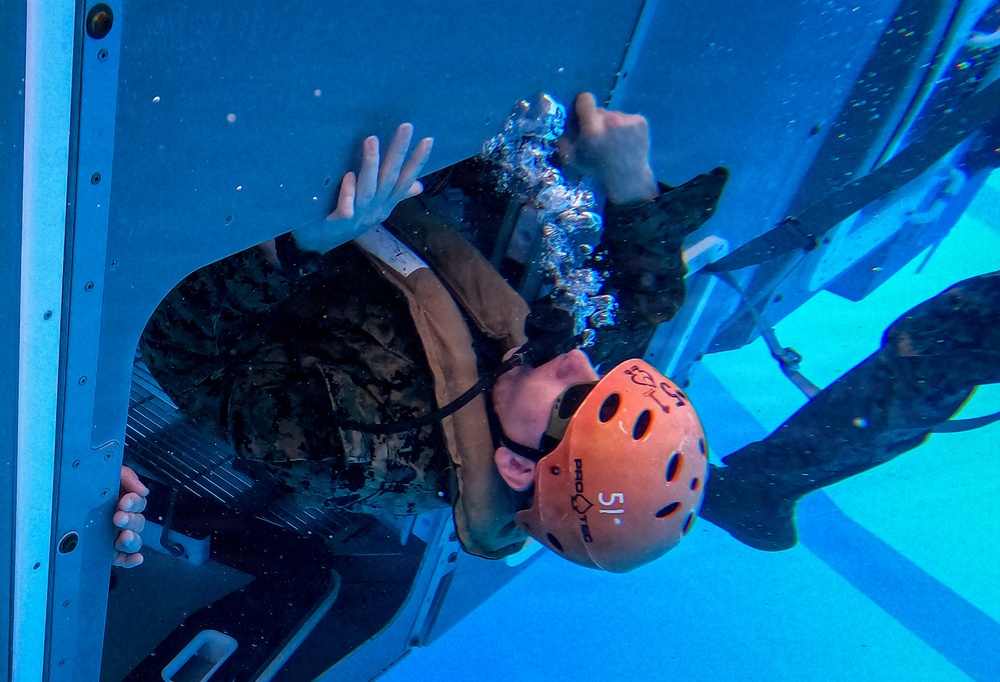 An individual wearing military camouflage uniform and an orange Pro-Tec helmet marked '51' is submerged underwater, appearing to exit or navigate through a hatch or window opening of a simulated vehicle or aircraft fuselage. The scene is consistent with Underwater Egress Training (UWET), a military survival exercise where personnel practice escaping from submerged vehicles. The image is taken from an underwater perspective, showing air bubbles rising around the subject.