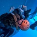 An individual wearing military camouflage uniform and an orange Pro-Tec helmet marked '51' is submerged underwater, appearing to exit or navigate through a hatch or window opening of a simulated vehicle or aircraft fuselage. The scene is consistent with Underwater Egress Training (UWET), a military survival exercise where personnel practice escaping from submerged vehicles. The image is taken from an underwater perspective, showing air bubbles rising around the subject.