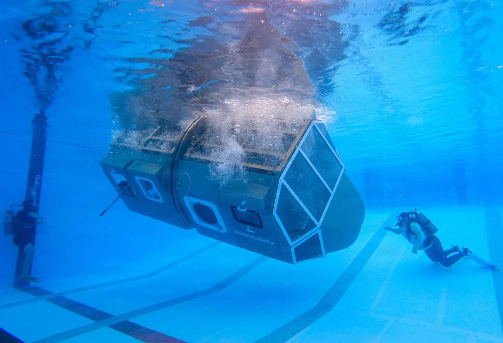 An underwater photograph taken inside a large pool showing a submersible or underwater habitat module being tested or deployed. A scuba diver is visible to the right, observing or monitoring the vessel. Air bubbles are escaping from the top of the structure, suggesting active pressurization or flooding procedures.