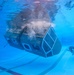 An underwater photograph taken inside a large pool showing a submersible or underwater habitat module being tested or deployed. A scuba diver is visible to the right, observing or monitoring the vessel. Air bubbles are escaping from the top of the structure, suggesting active pressurization or flooding procedures.