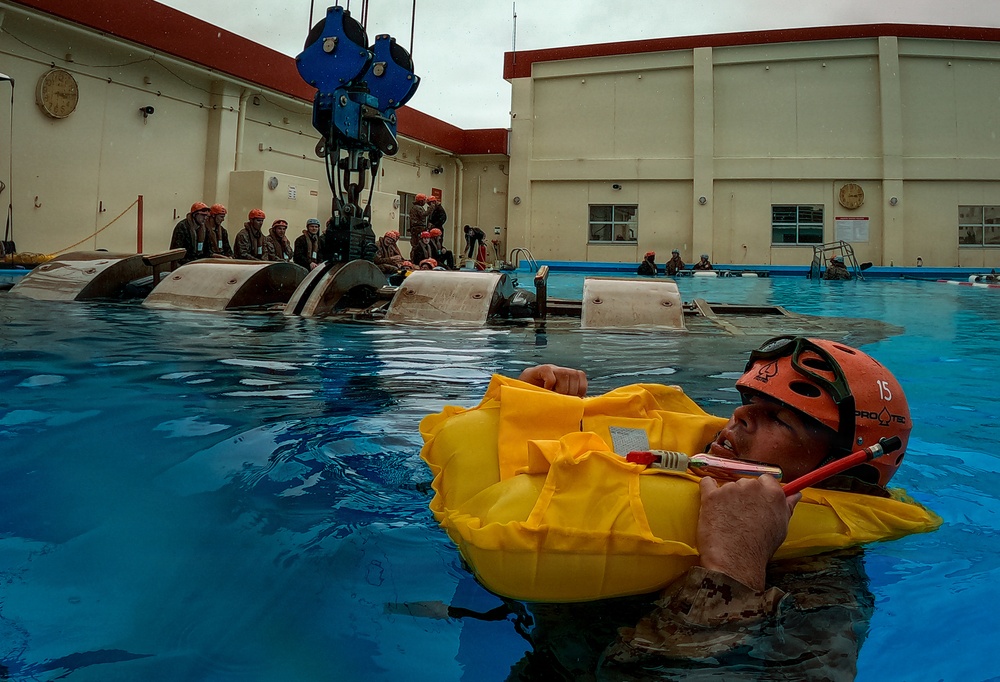 A military or aviation survival training exercise is taking place in an outdoor pool facility. A trainee wearing a red helmet marked '15' and an inflated yellow life preserver is floating in the water, while a submerged or partially inverted helicopter/aircraft fuselage simulator is visible in the background. Multiple other trainees in matching red helmets observe from the sides of the pool.