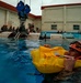 A military or aviation survival training exercise is taking place in an outdoor pool facility. A trainee wearing a red helmet marked '15' and an inflated yellow life preserver is floating in the water, while a submerged or partially inverted helicopter/aircraft fuselage simulator is visible in the background. Multiple other trainees in matching red helmets observe from the sides of the pool.
