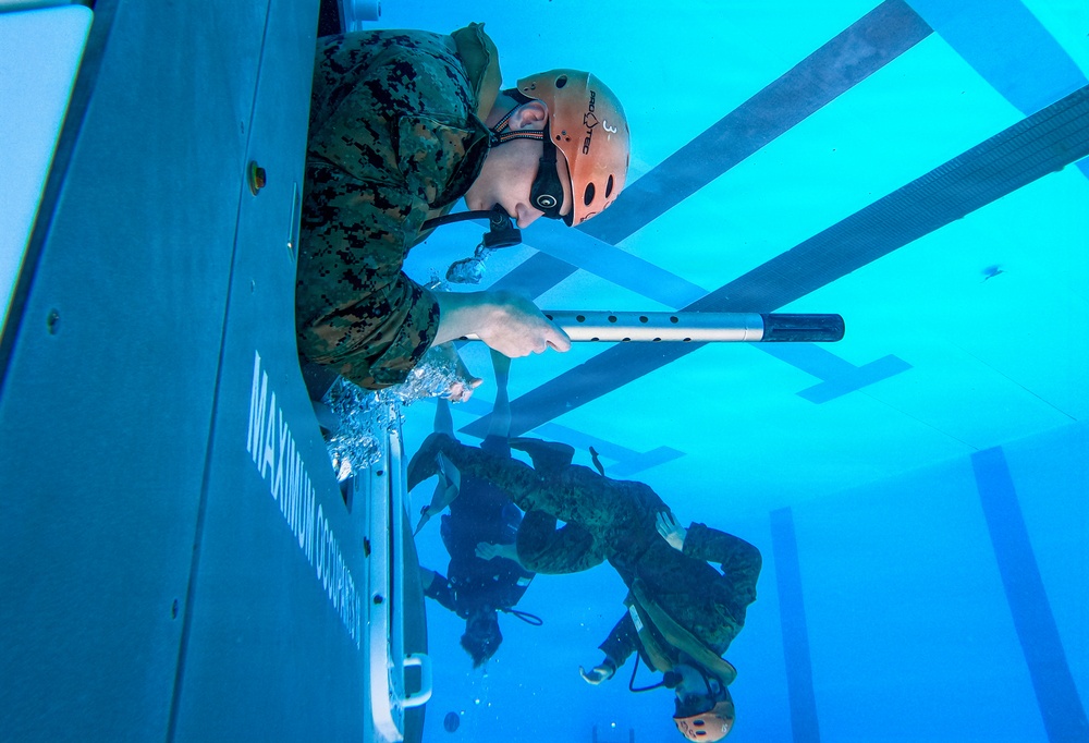 Military personnel, appearing to be U.S. Marines based on their camouflage uniforms, are conducting underwater egress or helicopter dunker training in a pool facility. The photo is taken from an underwater perspective, showing one Marine in the foreground gripping a bar while exiting a submerged simulated aircraft fuselage, with additional personnel visible underwater in the background. This type of training prepares service members to escape from submerged aircraft or vehicles.