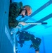 Military personnel, appearing to be U.S. Marines based on their camouflage uniforms, are conducting underwater egress or helicopter dunker training in a pool facility. The photo is taken from an underwater perspective, showing one Marine in the foreground gripping a bar while exiting a submerged simulated aircraft fuselage, with additional personnel visible underwater in the background. This type of training prepares service members to escape from submerged aircraft or vehicles.