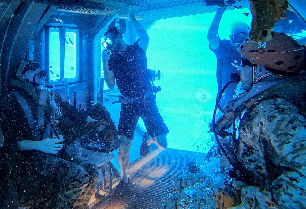 The image shows personnel inside what appears to be a submerged or partially flooded aircraft/helicopter cabin, likely during an underwater egress training exercise conducted in a pool or controlled water environment. The interior is illuminated by bright blue light from the water outside the open door/hatch. At least three individuals are visible, with one in civilian dive attire and others in military uniforms and helmets.