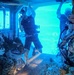 The image shows personnel inside what appears to be a submerged or partially flooded aircraft/helicopter cabin, likely during an underwater egress training exercise conducted in a pool or controlled water environment. The interior is illuminated by bright blue light from the water outside the open door/hatch. At least three individuals are visible, with one in civilian dive attire and others in military uniforms and helmets.