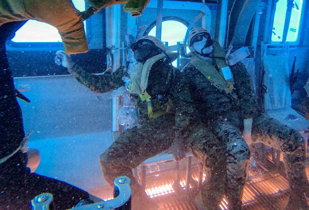 Two military personnel in camouflage uniforms and helmets with oxygen/breathing masks are positioned at what appears to be an open aircraft door or hatch, likely preparing for or executing a jump. The image is taken from inside the aircraft looking outward, with water spray or precipitation visible. The scene suggests a military airborne or water insertion training exercise.