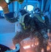 Two military personnel in camouflage uniforms and helmets with oxygen/breathing masks are positioned at what appears to be an open aircraft door or hatch, likely preparing for or executing a jump. The image is taken from inside the aircraft looking outward, with water spray or precipitation visible. The scene suggests a military airborne or water insertion training exercise.