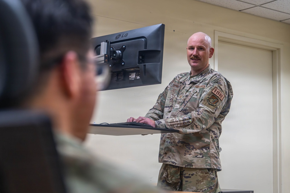 A U.S. Air Force service member in OCP (Operational Camouflage Pattern) uniform stands at a wall-mounted workstation with an LG monitor and keyboard. The individual appears to be in a briefing or instructional setting, facing toward another person in the foreground. An 'ISR' patch is visible on the right sleeve, indicating an Intelligence, Surveillance, and Reconnaissance role.