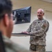 A U.S. Air Force service member in OCP (Operational Camouflage Pattern) uniform stands at a wall-mounted workstation with an LG monitor and keyboard. The individual appears to be in a briefing or instructional setting, facing toward another person in the foreground. An 'ISR' patch is visible on the right sleeve, indicating an Intelligence, Surveillance, and Reconnaissance role.