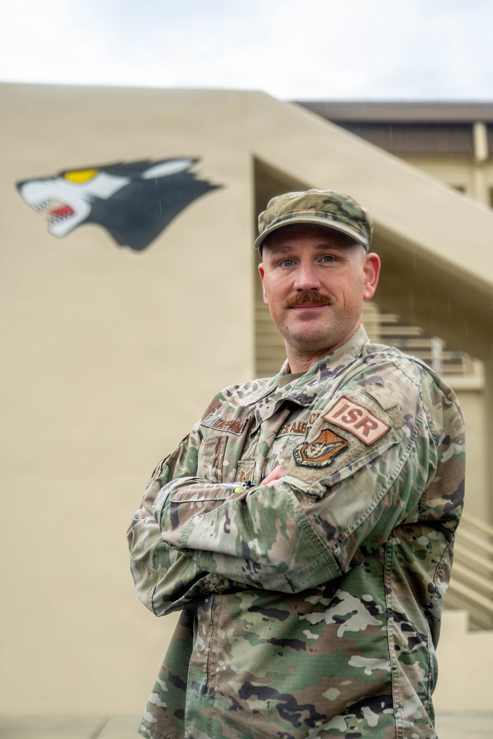 A U.S. Air Force service member in OCP (Operational Camouflage Pattern) uniform stands with arms crossed in front of a tan military building. The individual wears an 'ISR' patch on the right sleeve and a unit patch on the left sleeve, indicating an Intelligence, Surveillance, and Reconnaissance assignment. A wolf or panther mascot mural is painted on the building wall behind them.