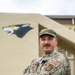 A U.S. Air Force service member in OCP (Operational Camouflage Pattern) uniform stands with arms crossed in front of a tan military building. The individual wears an 'ISR' patch on the right sleeve and a unit patch on the left sleeve, indicating an Intelligence, Surveillance, and Reconnaissance assignment. A wolf or panther mascot mural is painted on the building wall behind them.