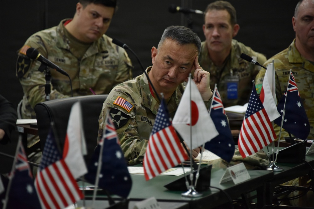 A multinational military meeting or conference is taking place, with personnel from the United States, Japan, and Australia present at a conference table. Small national flags representing the USA, Japan, and Australia are arranged on the table, indicating a trilateral or joint military coordination session. The central figure, wearing a U.S. Army uniform with a 2nd Infantry Division patch and a name tape reading 'DOOLAN,' appears to be in focused contemplation.