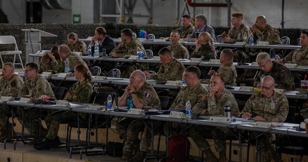 A multinational military briefing or conference is taking place in what appears to be a large hangar or military facility. Multiple military personnel in various camouflage uniforms are seated at rows of tables with name placards, water bottles, and documents. At least one French military officer is visible, suggesting a coalition or NATO-type gathering.