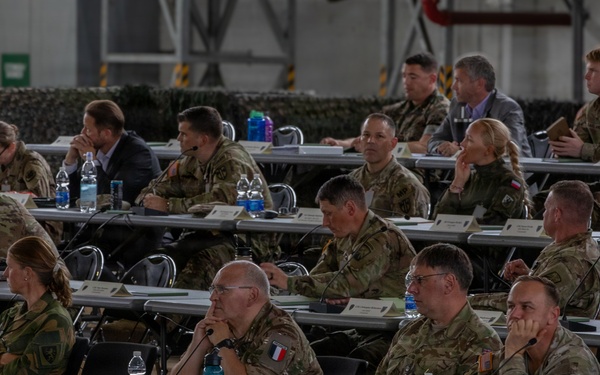 A multinational military briefing or conference is taking place in what appears to be a large hangar or military facility. Multiple military personnel in various camouflage uniforms are seated at rows of tables with name placards, water bottles, and documents. At least one French military officer is visible, suggesting a coalition or NATO-type gathering.