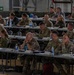A multinational military briefing or conference is taking place in what appears to be a large hangar or military facility. Multiple military personnel in various camouflage uniforms are seated at rows of tables with name placards, water bottles, and documents. At least one French military officer is visible, suggesting a coalition or NATO-type gathering.