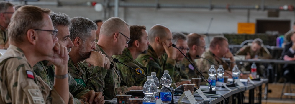 A multinational military briefing or conference is taking place, with numerous uniformed personnel seated along a long table in what appears to be a hangar or large military facility. Personnel are wearing various national military uniforms, including at least one Polish flag patch visible on the leftmost individual. The attendees appear to be attentively listening to a presentation or briefing.